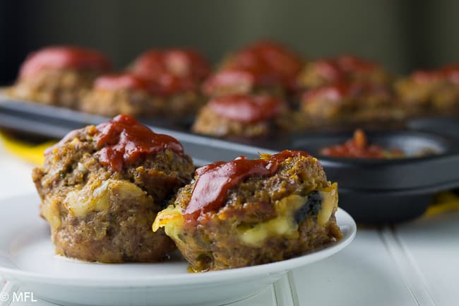 mozzarella stuffed meatloaf covered in ketchup on white plate with cupcake pan in back with meatloaves in it