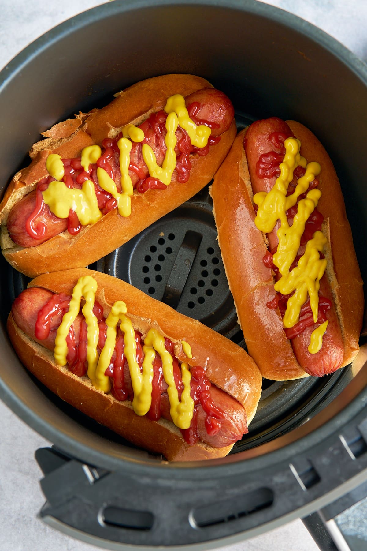 hot dogs in buns in air fryer basket topped with ketchup and mustard