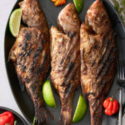 A black serving platter topped with 3 whole snapper fish in a Jamaican jerk marinade garnished with Scotch Bonnet peppers, wedges of lime and sprigs of thyme.