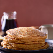 A stack of sweet potato pancakes on a black plate with a jug of maple syrup served alongside.