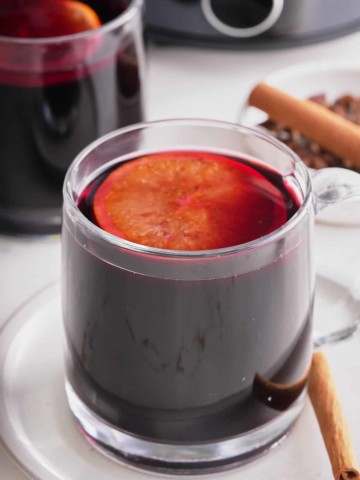 Two glass cups filled with mulled wine and topped with a slice of mulled orange with whole spices set alongside.