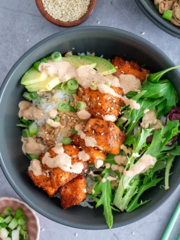 salmon bowl on table with cooked salmon, rice, greens, topping and sauce in bowl