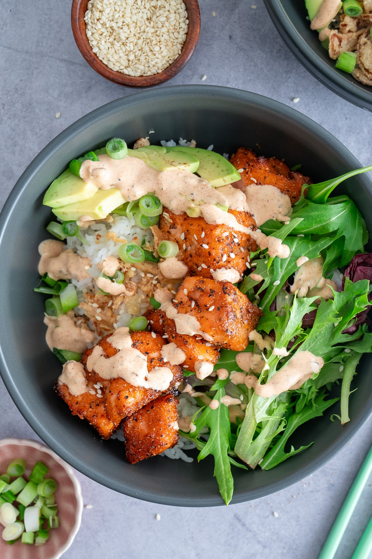 salmon bowl on table with cooked salmon, rice, greens, topping and sauce in bowl